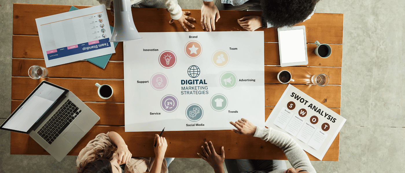 Overhead view of a team collaborating around a table, pointing at a large printout of a 'Digital Marketing Strategies' diagram.