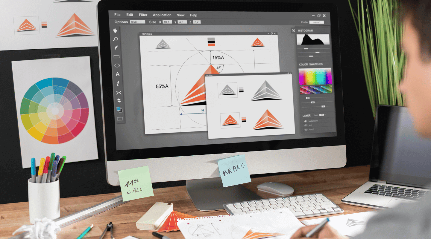 Graphic designer creating a logo, displaying vector measurements and color palettes on a monitor.