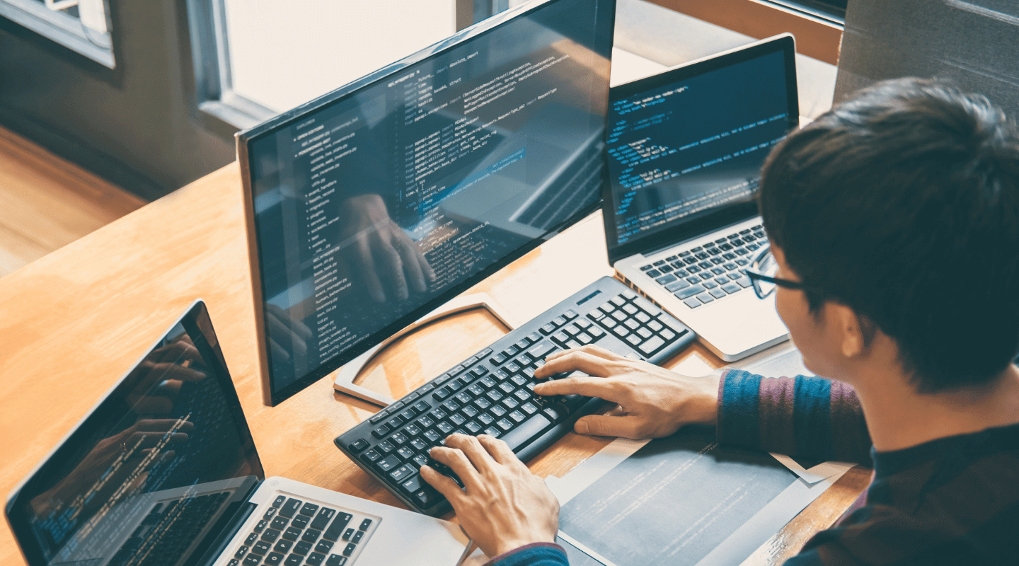 A person coding at a desk with a large central monitor and two flanking laptops showing lines of software code.